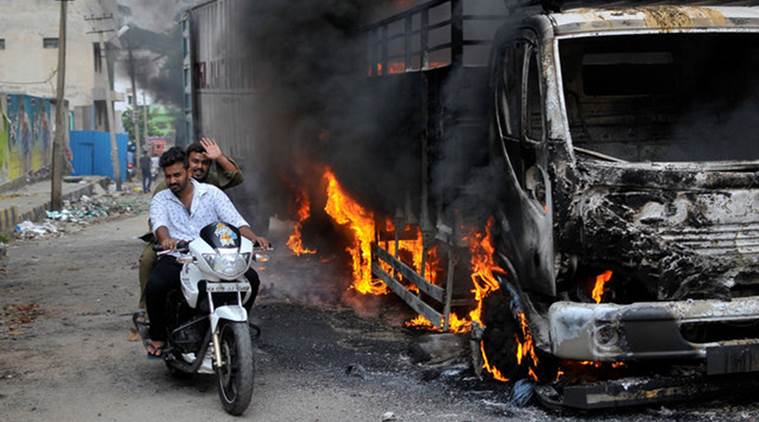 Men ride a motorcycle past a lorry which was set on fire by protesters in Bengaluru