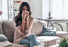 Cold and flu. Sick young woman blowing the nose using tissue paper while sitting on the sofa at home