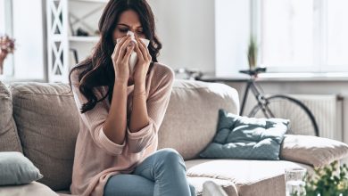 Cold and flu. Sick young woman blowing the nose using tissue paper while sitting on the sofa at home