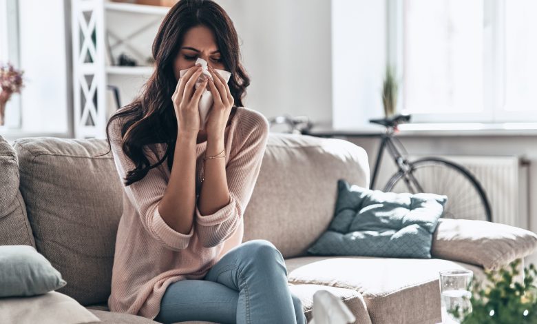 Cold and flu. Sick young woman blowing the nose using tissue paper while sitting on the sofa at home
